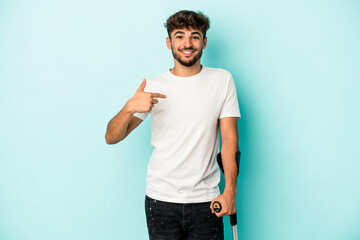 Young arab man with crutches isolated on blue background person pointing by hand to a shirt copy space, proud and confident