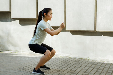 Positive Asian sportswoman with long dark ponytail does squats at training near stone steps and concrete wall on city street