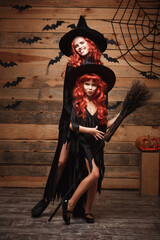 Fototapeta na wymiar Halloween Concept - Beautiful caucasian mother and her daughter with long red hair in witch costumes celebrating Halloween posing with over bats and spider web on Wooden studio background.