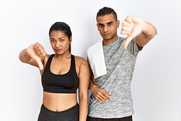 Fototapeta na wymiar Young latin couple wearing sportswear standing over isolated background looking unhappy and angry showing rejection and negative with thumbs down gesture. bad expression.