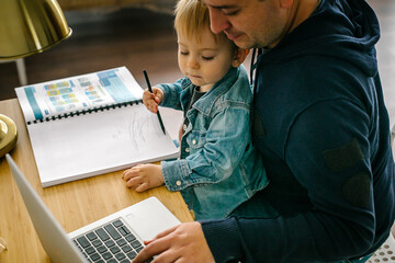 Father holding little baby son and working on laptop at home. Concept of remotely work.