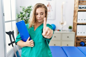 Young caucasian woman working at pain recovery clinic pointing with finger up and angry expression, showing no gesture
