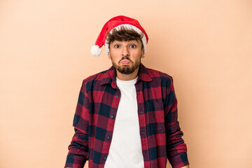 Young arab man celebrating Christmas isolated on beige background shrugs shoulders and open eyes confused.