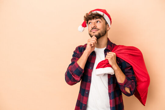 Young arab man disguised as santa claus isolated on beige background looking sideways with doubtful and skeptical expression.
