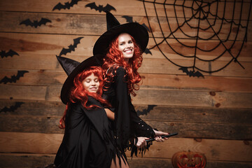 Fototapeta na wymiar Halloween Concept - Beautiful caucasian mother and her daughter with long red hair in witch costumes flying with magic broomstick.