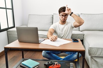 Hispanic man doing papers at home making fun of people with fingers on forehead doing loser gesture...