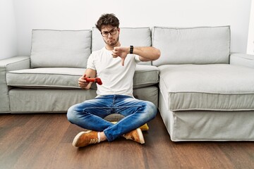 Hispanic man playing video game holding controller by the sofa with angry face, negative sign...