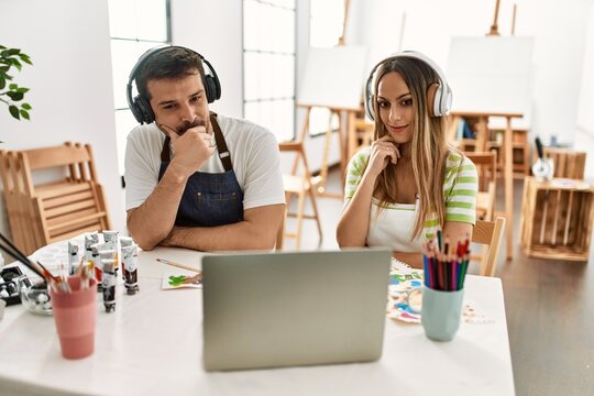 Young couple of wife and husband at art studio looking at video on laptop serious face thinking about question with hand on chin, thoughtful about confusing idea