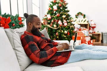 Young african american man using laptop sitting by christmas tree at home