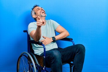 Handsome middle age man with grey hair sitting on wheelchair laughing at you, pointing finger to the camera with hand over body, shame expression