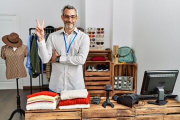Middle age hispanic man working as manager at retail boutique smiling with happy face winking at the camera doing victory sign with fingers. number two.