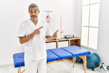 Middle age hispanic therapist man working at pain recovery clinic pointing aside worried and nervous with both hands, concerned and surprised expression