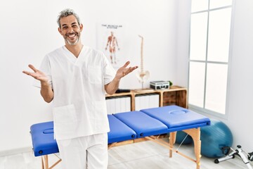 Middle age hispanic therapist man working at pain recovery clinic smiling cheerful offering hands giving assistance and acceptance.