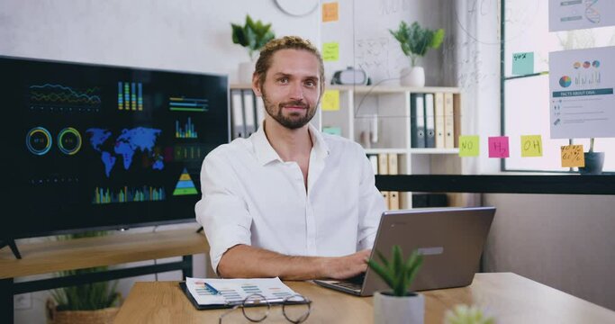 Portrait of handsome positive confident creative skilful bearded office worker which typing on computer in workroom and looking into camera with sincerely smile