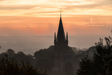 Obraz premium A typical colorful Autumn sunrise in Maastricht with the landscape covered with a layer of fog, leaving only silhouettes visible in the distance, like this tower of a church on the hillside.