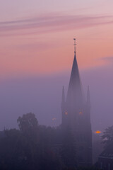 Fototapeta premium A typical colorful Autumn sunrise in Maastricht with the landscape covered with a layer of fog, leaving only silhouettes visible in the distance, like this tower of a church on the hillside.
