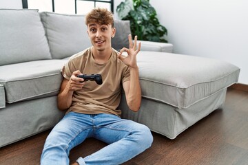 Young caucasian man playing video game holding controller at home doing ok sign with fingers, smiling friendly gesturing excellent symbol