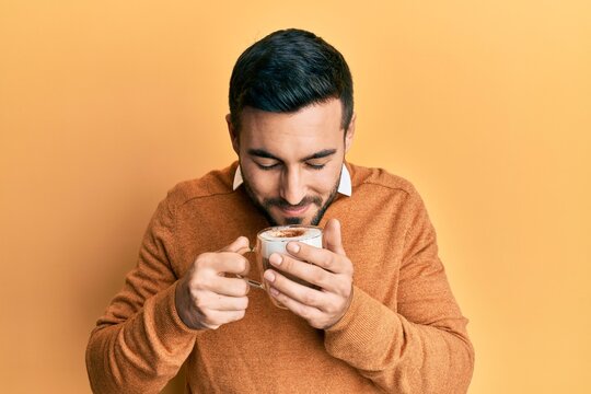 Handsome hispanic man enjoying a cup of coffee over yellow background