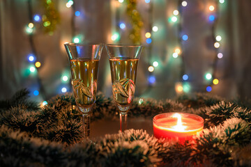 Fototapeta na wymiar Two glasses of sparkling wine surrounded by green and white tinsel with a burning candle and a luminous garland on a New Year's background.