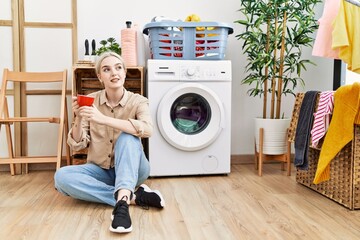 Young caucasian woman drinking coffee waiting for washing machine at laundry room