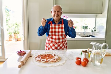 Senior man with grey hair cooking pizza at home kitchen very happy and excited doing winner gesture with arms raised, smiling and screaming for success. celebration concept.