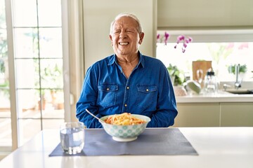 Senior man with grey hair eating pasta spaghetti at home with hand on stomach because nausea,...