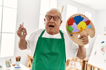 Senior grey-haired artist man surprised holding paintbrush and palette at art studio.