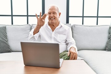 Senior man using laptop at home sitting on the sofa showing and pointing up with fingers number four while smiling confident and happy.