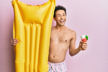 Obraz premium Young handsome man wearing swimsuit holding summer mattress float and icecream winking looking at the camera with sexy expression, cheerful and happy face.