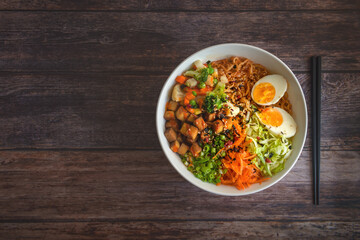 Variation of ramen homemade soup with noodles, marinated tofu, chopped carrots, onions, lettuce and broccoli in a bowl lying on a wooden table.