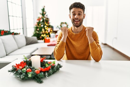 Arab young man sitting on the table by christmas tree celebrating surprised and amazed for success with arms raised and open eyes. winner concept.