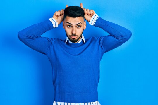 Young hispanic man with beard wearing casual blue sweater doing funny gesture with finger over head as bull horns