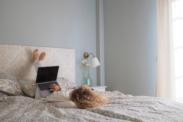 Young woman working on laptop while lying down on bed in bedroom. Businesswoman working from home while relaxing on bed. Lazy young woman typing on laptop and working in bedroom..