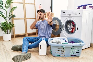 Young hispanic man putting dirty laundry into washing machine covering eyes with hands and doing stop gesture with sad and fear expression. embarrassed and negative concept.