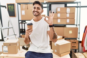 Young hispanic man working at small business ecommerce drinking a coffee doing ok sign with fingers, smiling friendly gesturing excellent symbol