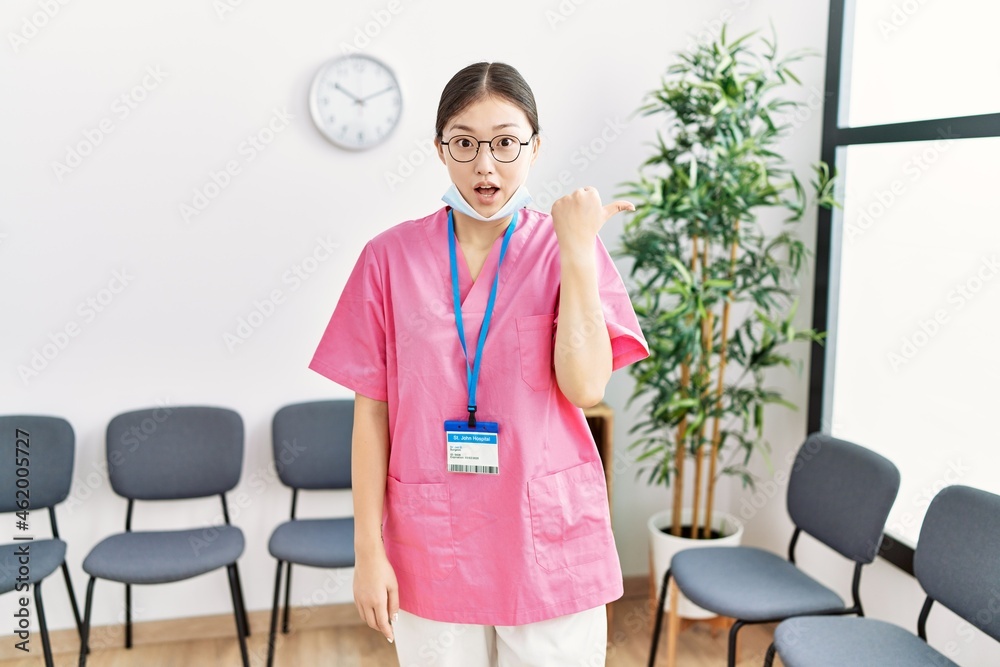 Sticker young asian nurse woman at medical waiting room surprised pointing with hand finger to the side, ope