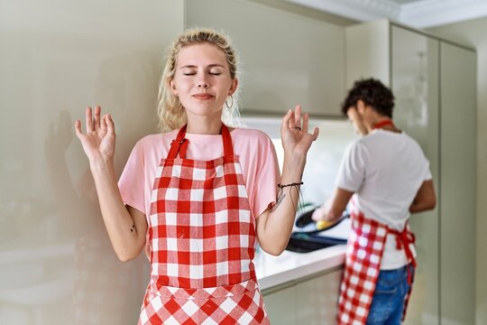 Young caucasian woman wearing apron and husband doing housework washing dishes relax and smiling with eyes closed doing meditation gesture with fingers. yoga concept.