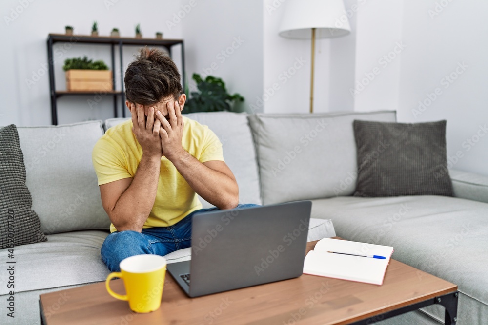 Poster Young man with beard using laptop at home with sad expression covering face with hands while crying. depression concept.