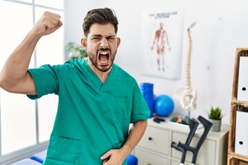 Young man with beard working at pain recovery clinic angry and mad raising fist frustrated and furious while shouting with anger. rage and aggressive concept.
