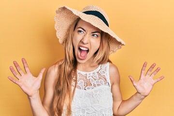 Young caucasian woman wearing summer hat crazy and mad shouting and yelling with aggressive expression and arms raised. frustration concept.