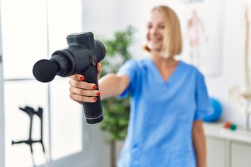 Young blonde woman wearing physiotherapist uniform holding massage pistol at rehab clinic