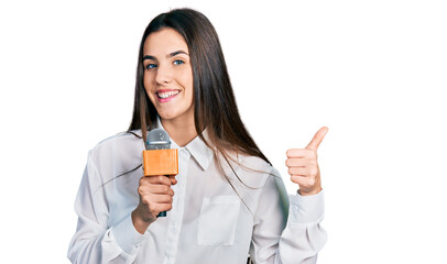 Young brunette teenager holding reporter microphone smiling happy and positive, thumb up doing excellent and approval sign