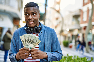 Young african american man using headphones holding dollars at the city.