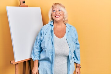 Middle age blonde woman standing by painter easel stand looking away to side with smile on face, natural expression. laughing confident.