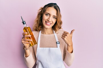 Young caucasian woman wearing apron holding olive oil can smiling happy and positive, thumb up...