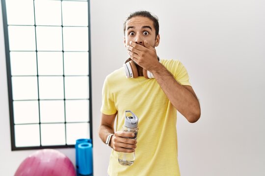 Young hispanic man wearing sportswear and drinking water at the gym shocked covering mouth with hands for mistake. secret concept.