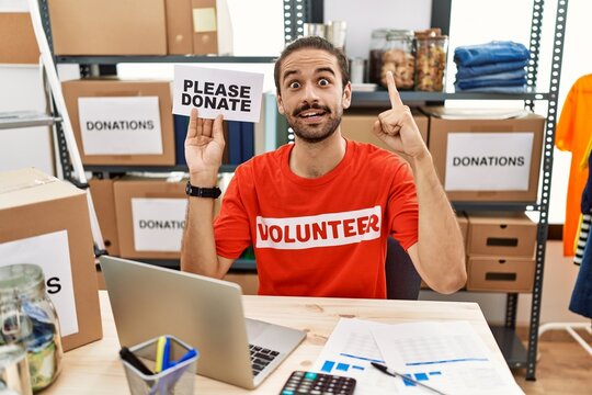 Young hispanic man wearing volunteer t shirt holding please donate banner smiling with an idea or question pointing finger with happy face, number one