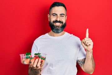 Young hispanic man holding strawberries smiling with an idea or question pointing finger with happy face, number one