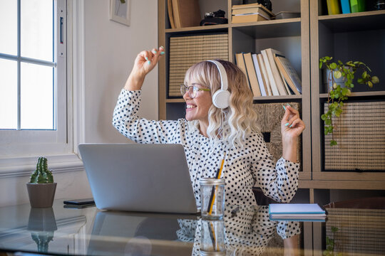 Businesswoman listening music on headphones while working on laptop at home office. Caucasian young woman enjoying music on headphones while looking out through window. Joyful woman with laptop 
