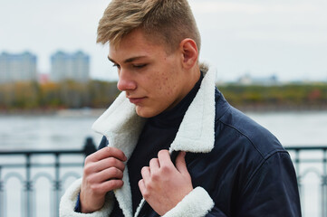 Portrait of a young Caucasian blond guy in a warm leather black jacket. The guy lifts the fur...